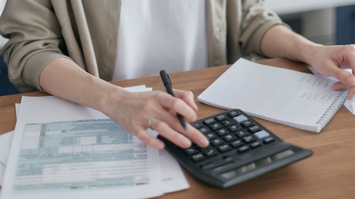 Mulher revisando cálculos e anotações financeiras em casa, com calculadora, caderno e papéis sobre a mesa